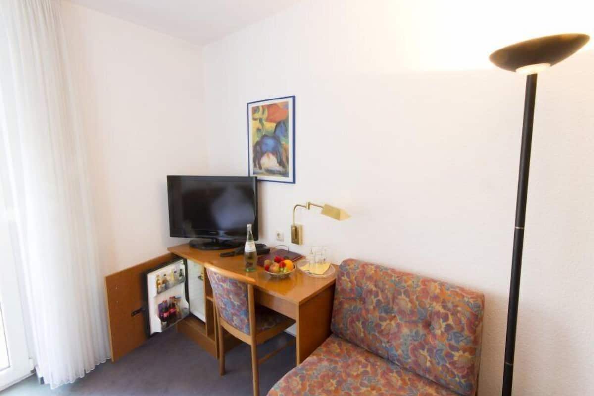 A hotel room with a TV, a desk with a lamp, a sofa, and a painting on the wall.