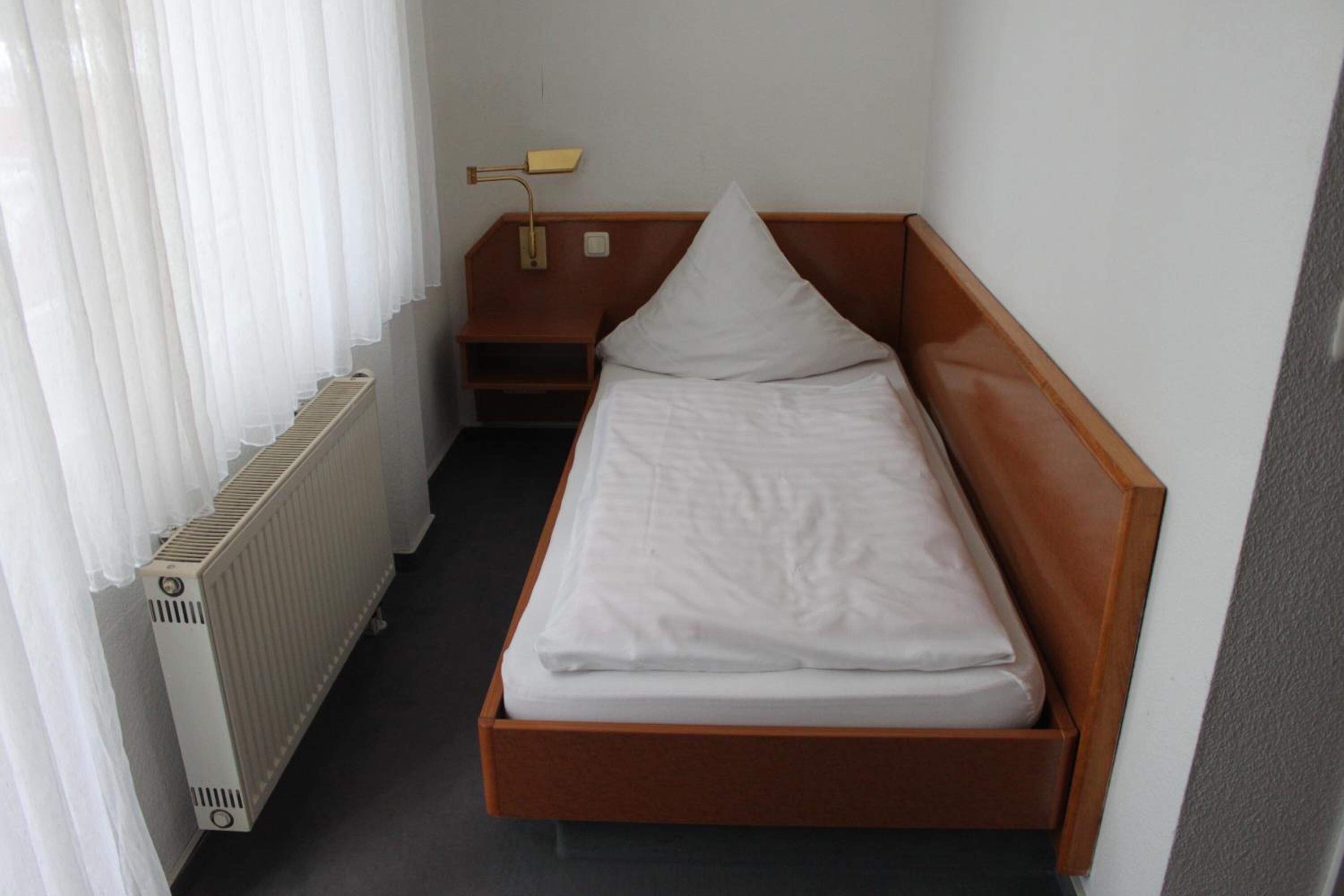Una habitación pequeña con cama individual, cabecera de madera, mesita de noche con lámpara y radiador.