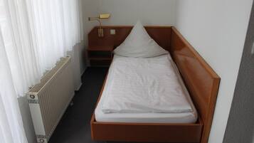 Una habitación pequeña con cama individual, cabecera de madera, mesita de noche con lámpara y radiador.