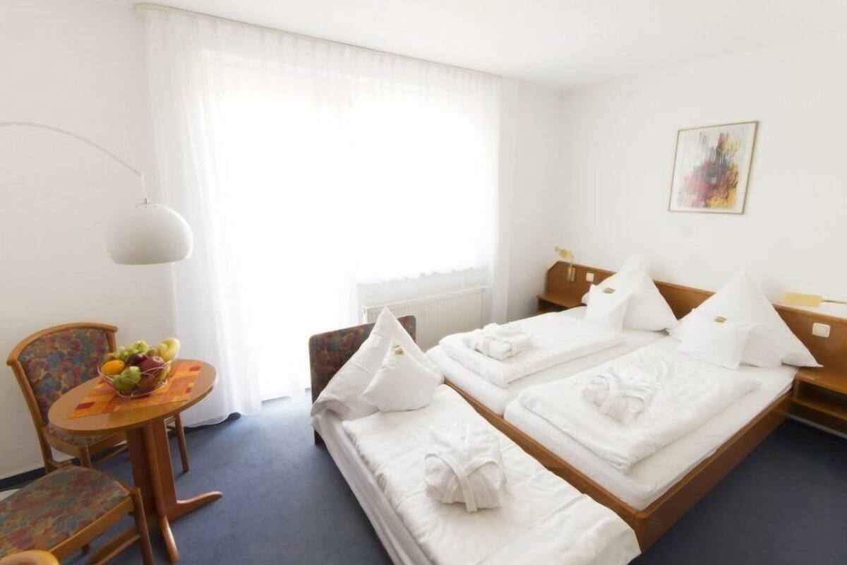 A hotel room with two beds, a small table with fruits, a chair, and a window with curtains.