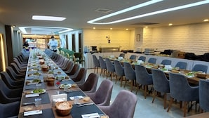Una larga mesa de comedor dispuesta con platos, cubiertos y cestas, bordeada por filas de sillas en un área de comedor moderna de un hotel.