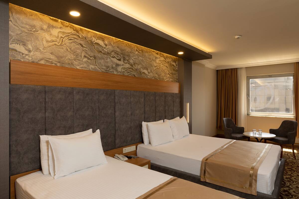 A hotel room with two beds, a marble headboard, a small table with chairs, and a window with curtains.