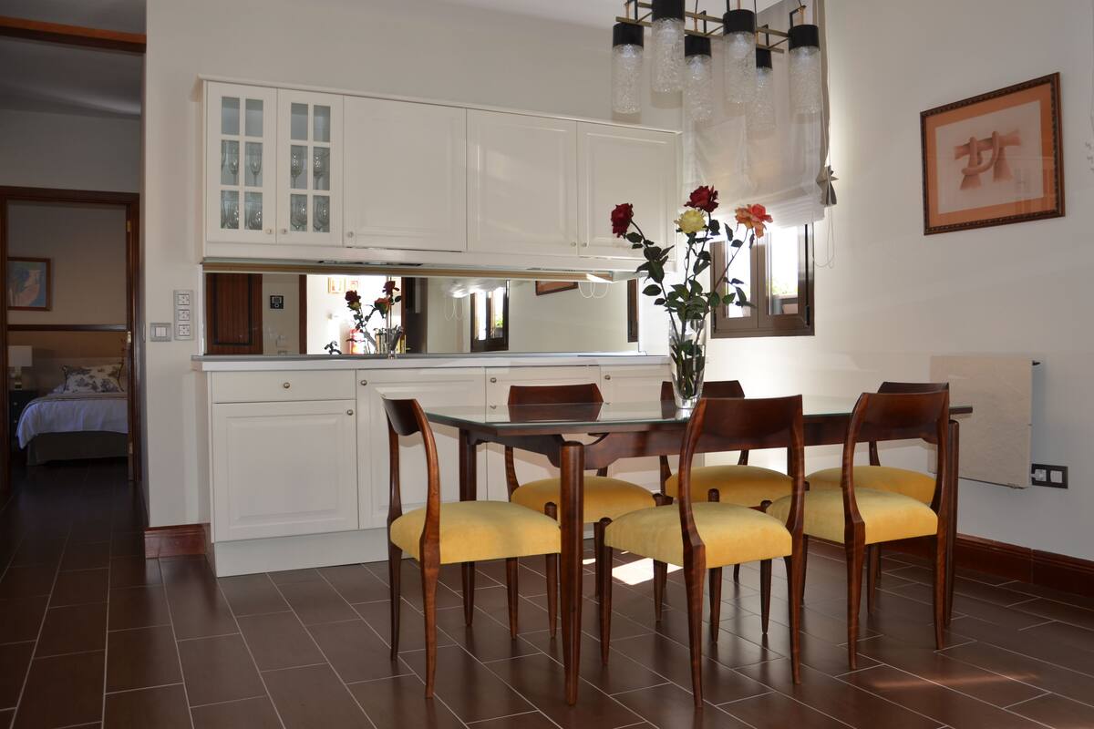 Uma área de jantar com uma mesa de vidro, cadeiras de madeira e um vaso com flores.