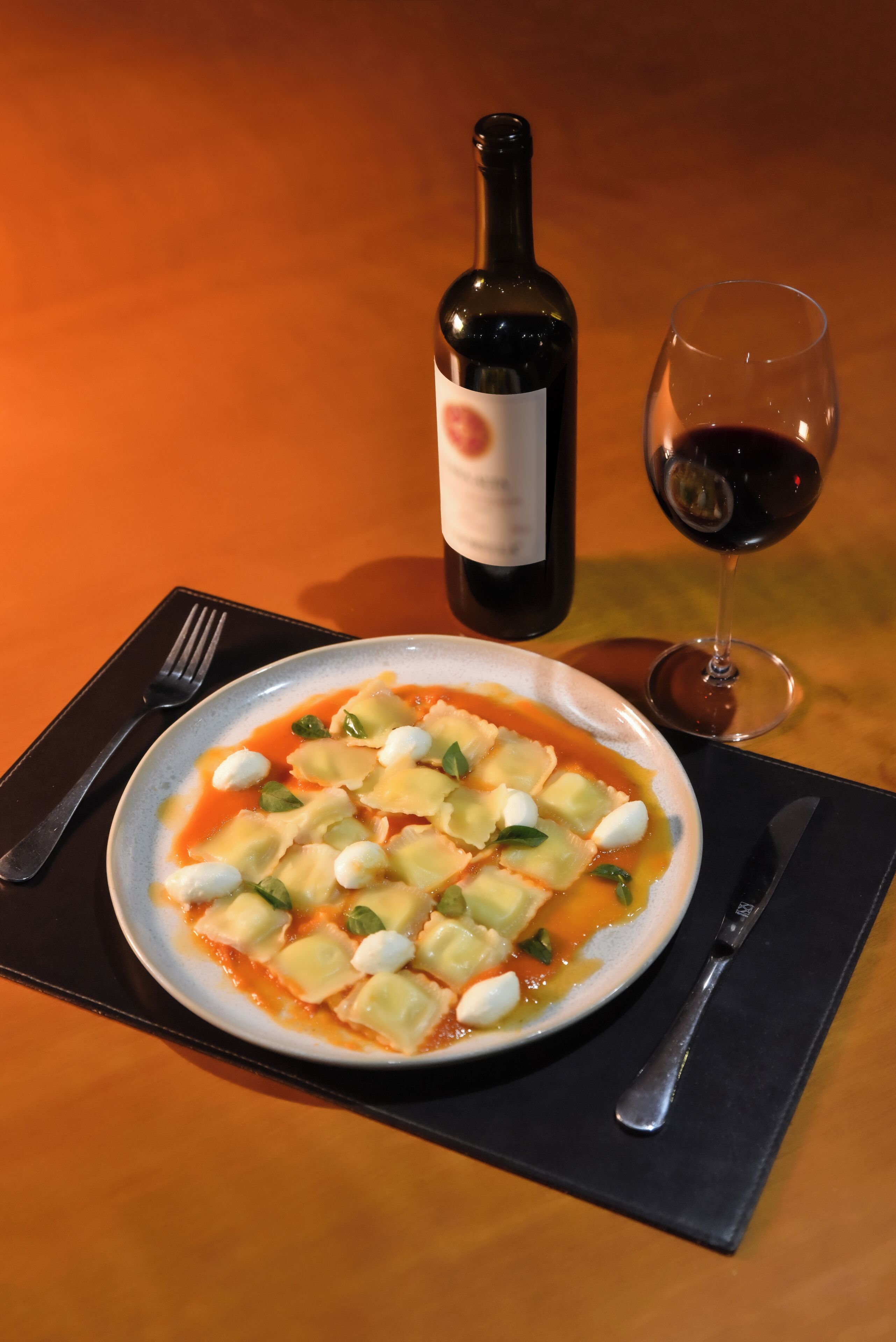 A plate of ravioli with vegetables and cheese, a bottle of wine, and a glass of red wine on a table.