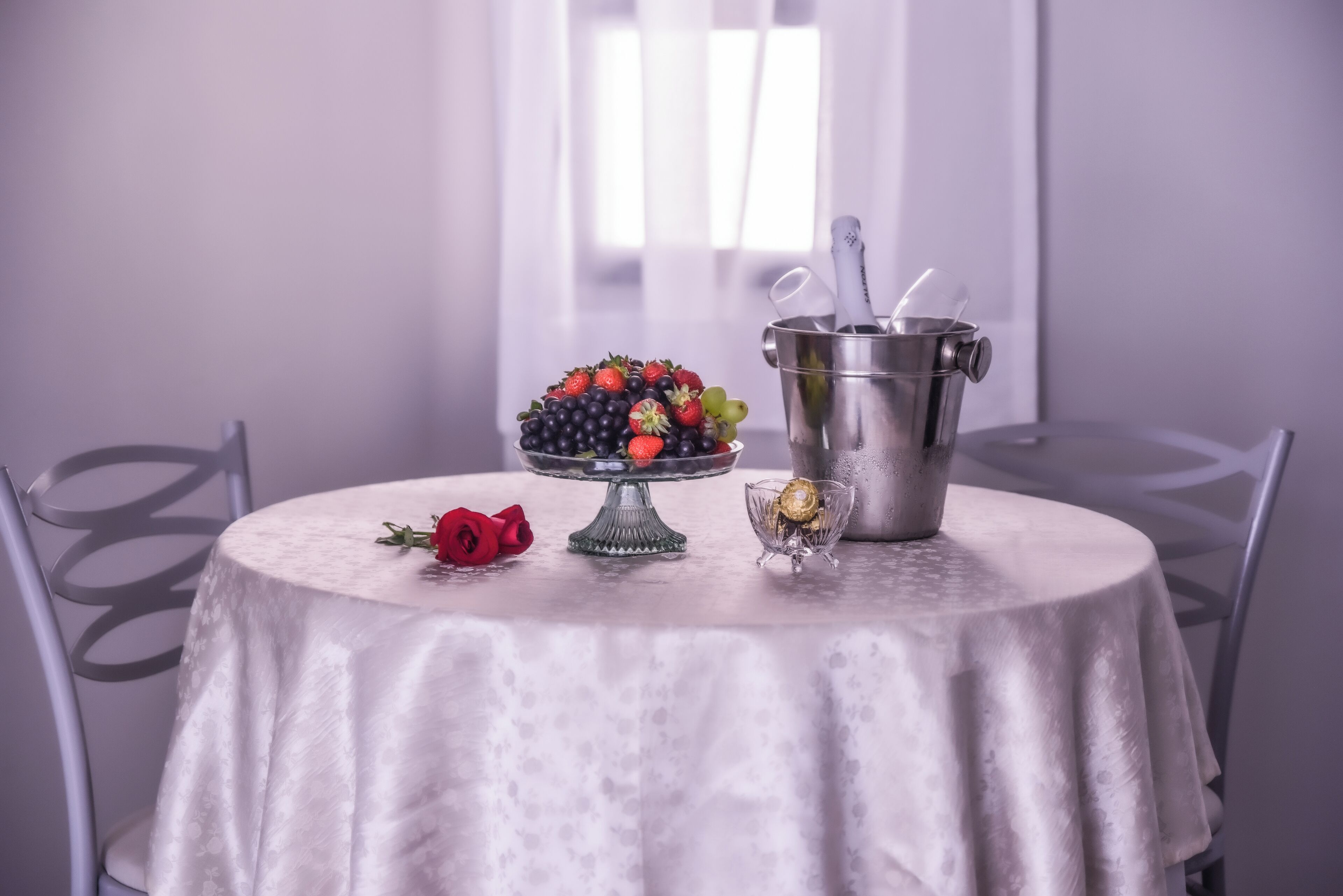 A round dining table with a white tablecloth, a fruit bowl with strawberries and grapes, a red rose, a glass of champagne, and a metal bucket with a spoon.