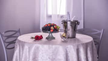 Une table de salle à manger ronde recouverte d’une nappe blanche, un panier de fruits contenant des fraises et des raisins, une rose rouge, un verre de champagne et un seau en métal avec une cuillère.
