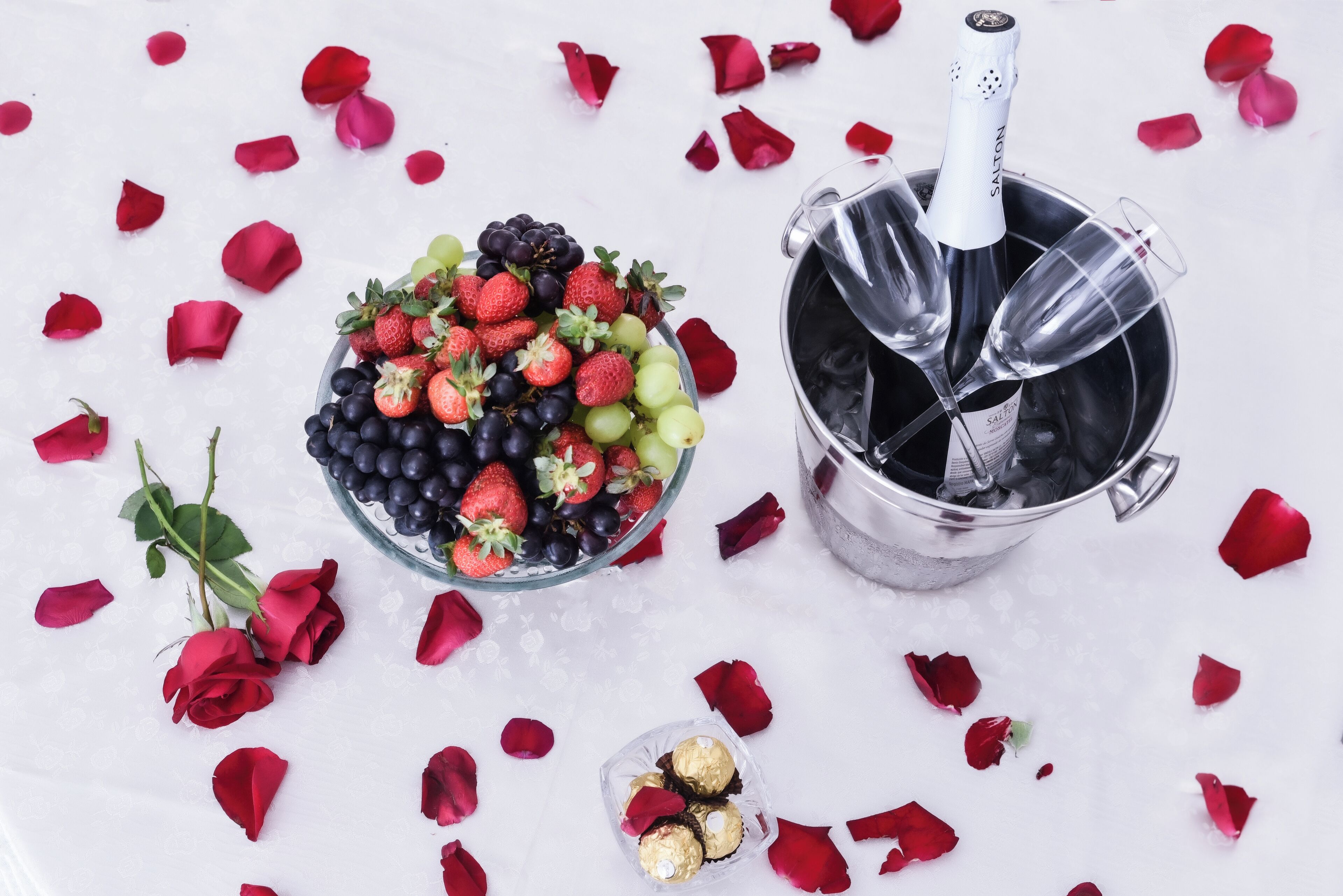 Una botella de champán, un cuenco de fruta y pétalos de rosa sobre una superficie blanca.
