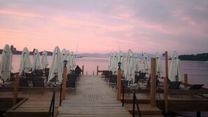 A wooden pier with rows of tables and chairs, some with white umbrellas, overlooking a calm body of water with boats in the distance.