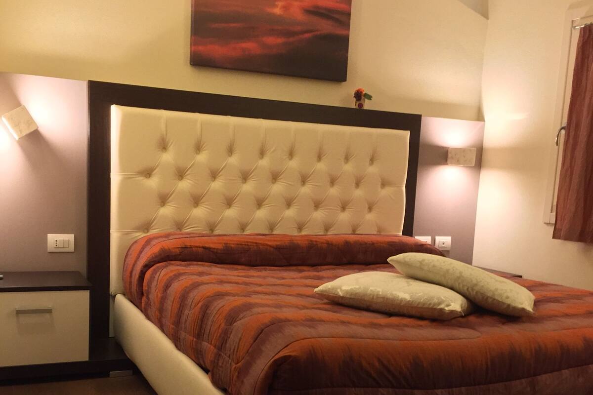 Une chambre à coucher avec une tête de lit capitonnée, un lit avec une literie rouge et blanche, et deux lampes de chevet.