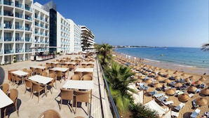 Un hotel frente a la playa con asientos al aire libre y sombrillas, una zona de piscina y vistas a la playa y al océano.