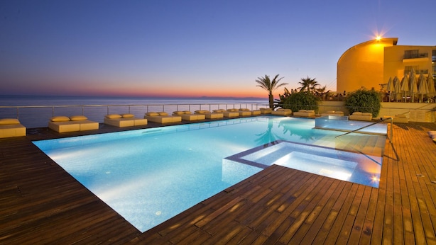 Zona de piscina con vista al océano al atardecer, terraza de madera y sillones de descanso.