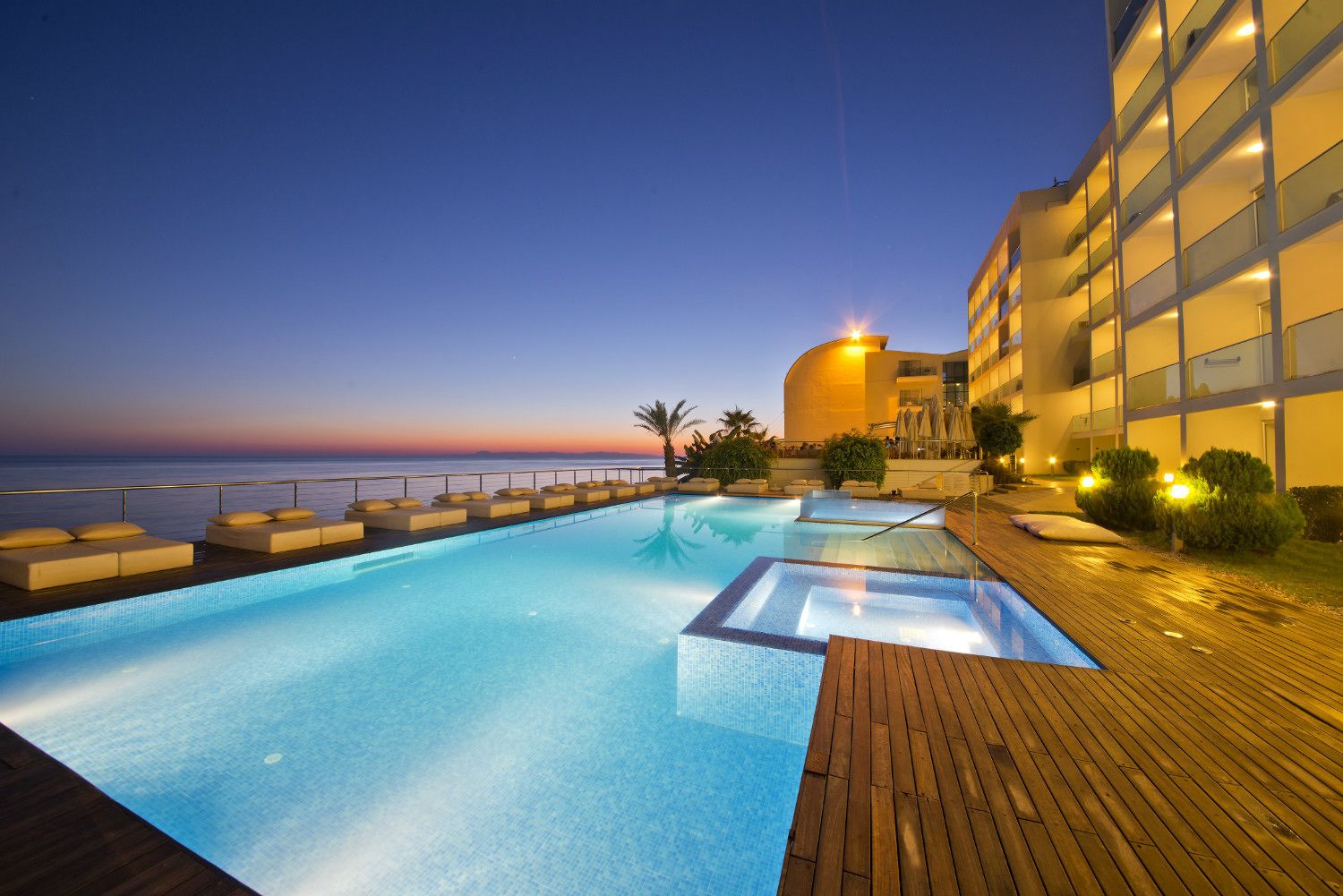 Une piscine dotée d’une terrasse en bois et de chaises longues, entourée d’un bâtiment à la façade incurvée, offrant une vue sur la mer au crépuscule.