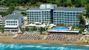 Un hotel frente a la playa con varias construcciones, un amplio espacio de piscina y numerosos sillones de sol en la arena.