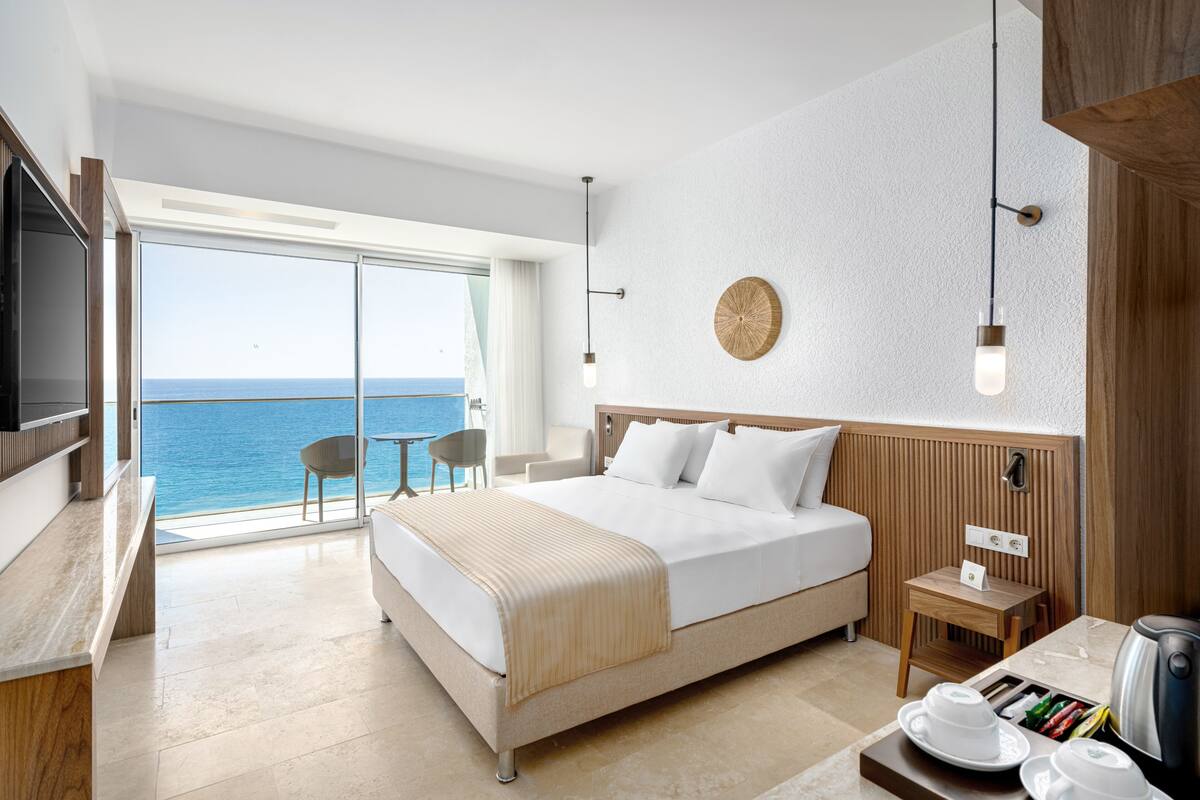 A modern hotel room with a large bed, a desk, a chair, and a view of the sea.