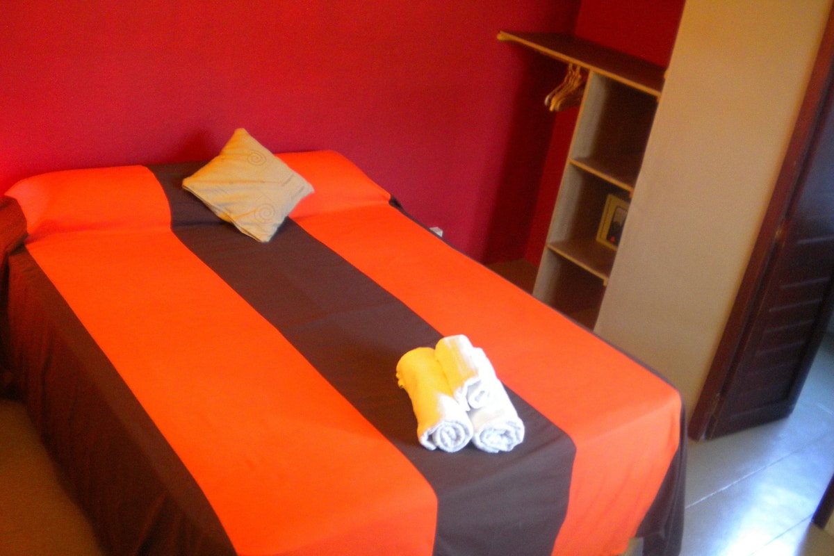 A single bed with an orange and brown bedspread, a pillow, and two rolled towels on the foot of the bed.