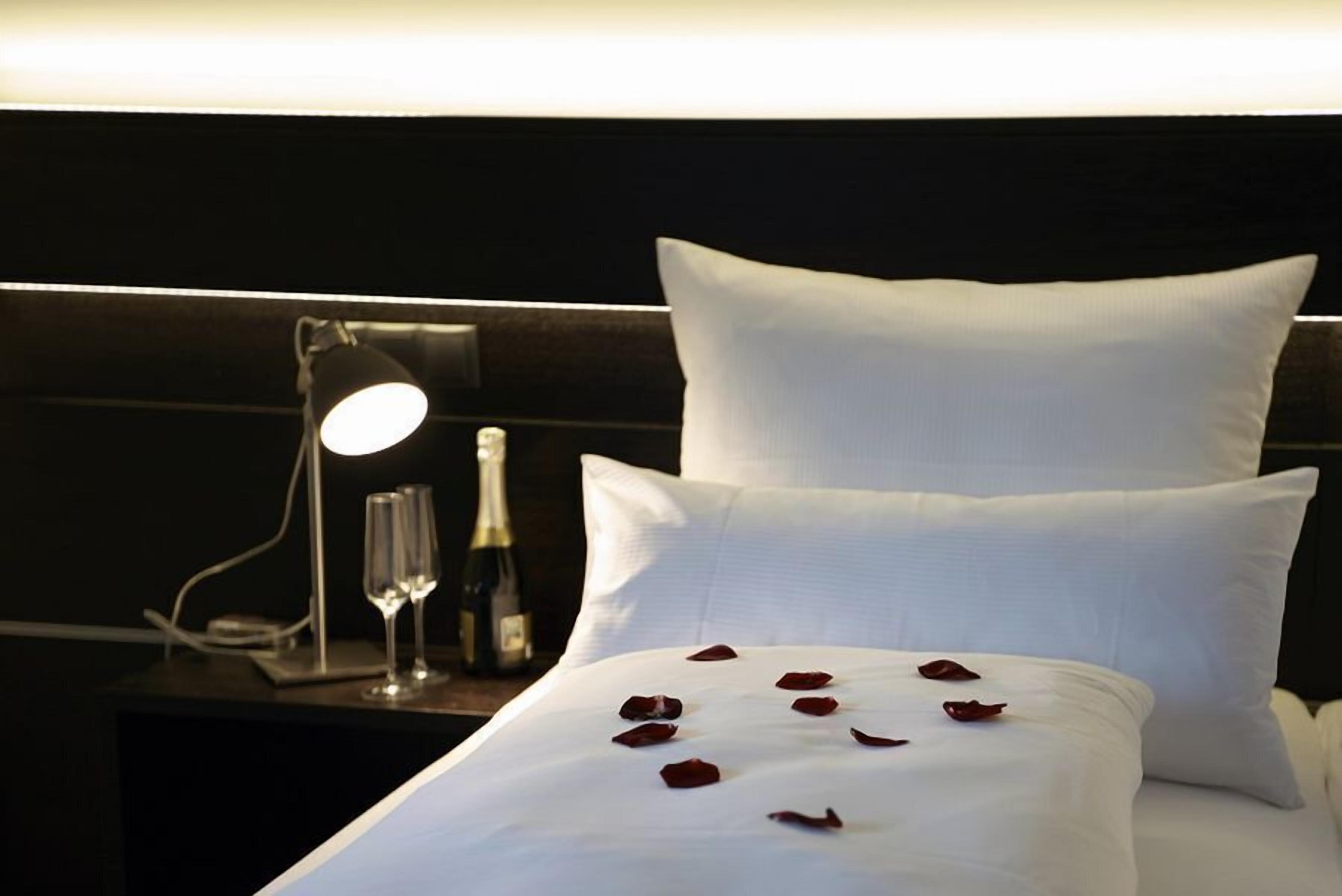 Ein ordentlich bezogenes Bett mit weißen Leinentüchern und Rosenblättern, eine Nachttischlampe und eine Champagnerflasche auf einem dunklen Holznachttisch.