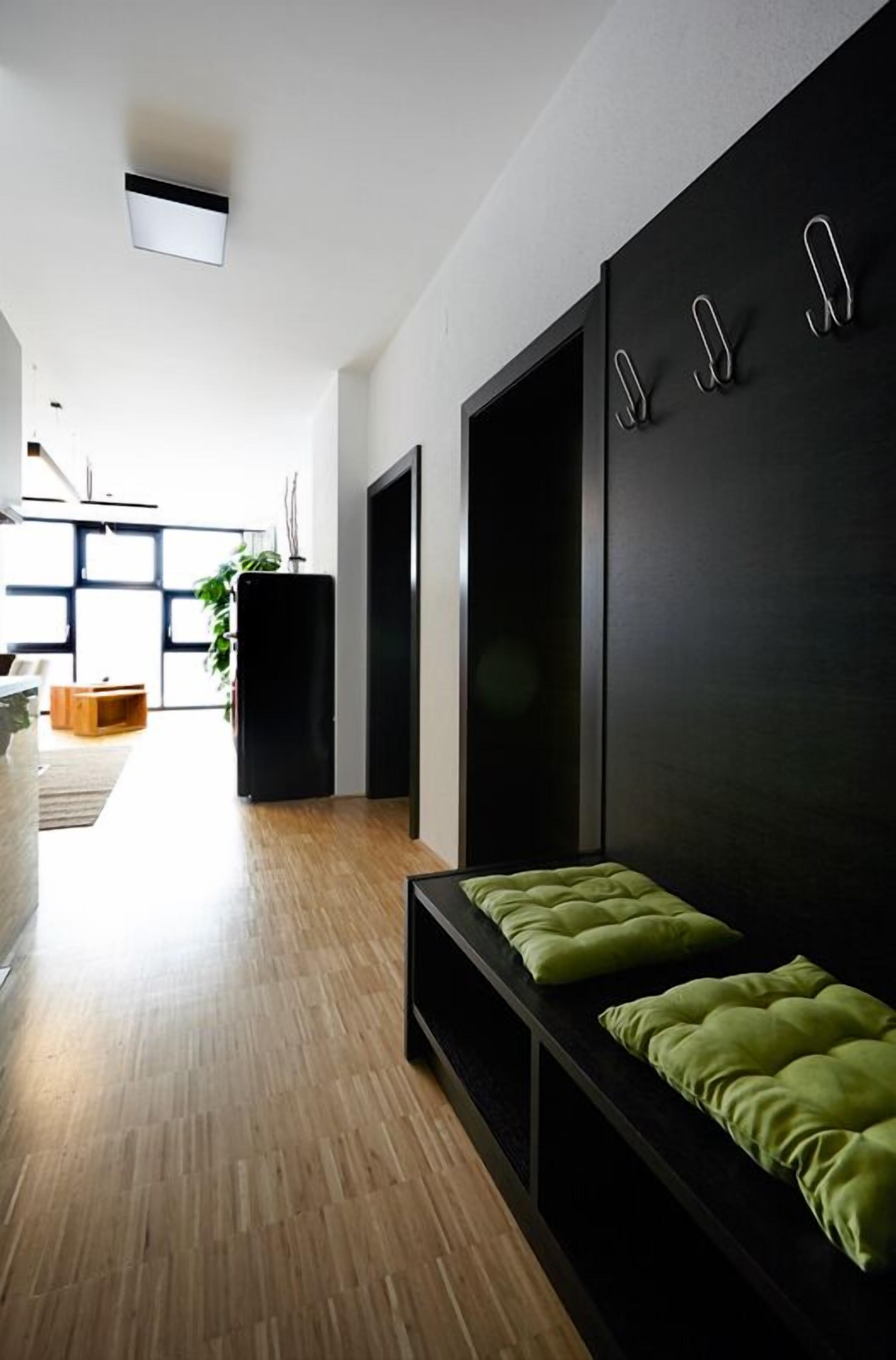 Un interior moderno con piso de madera, un sofá negro con cojines verdes y un estante montado en la pared con ganchos.