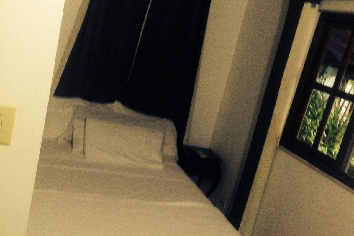 Une petite pièce faiblement éclairée, avec un lit simple, une table de chevet et une fenêtre donnant sur des arbres.