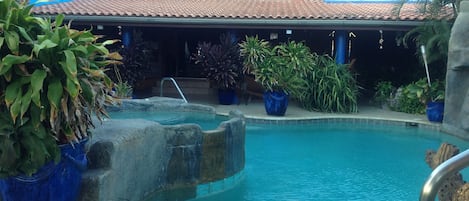 A pool area with a blue building in the background, surrounded by various potted plants.