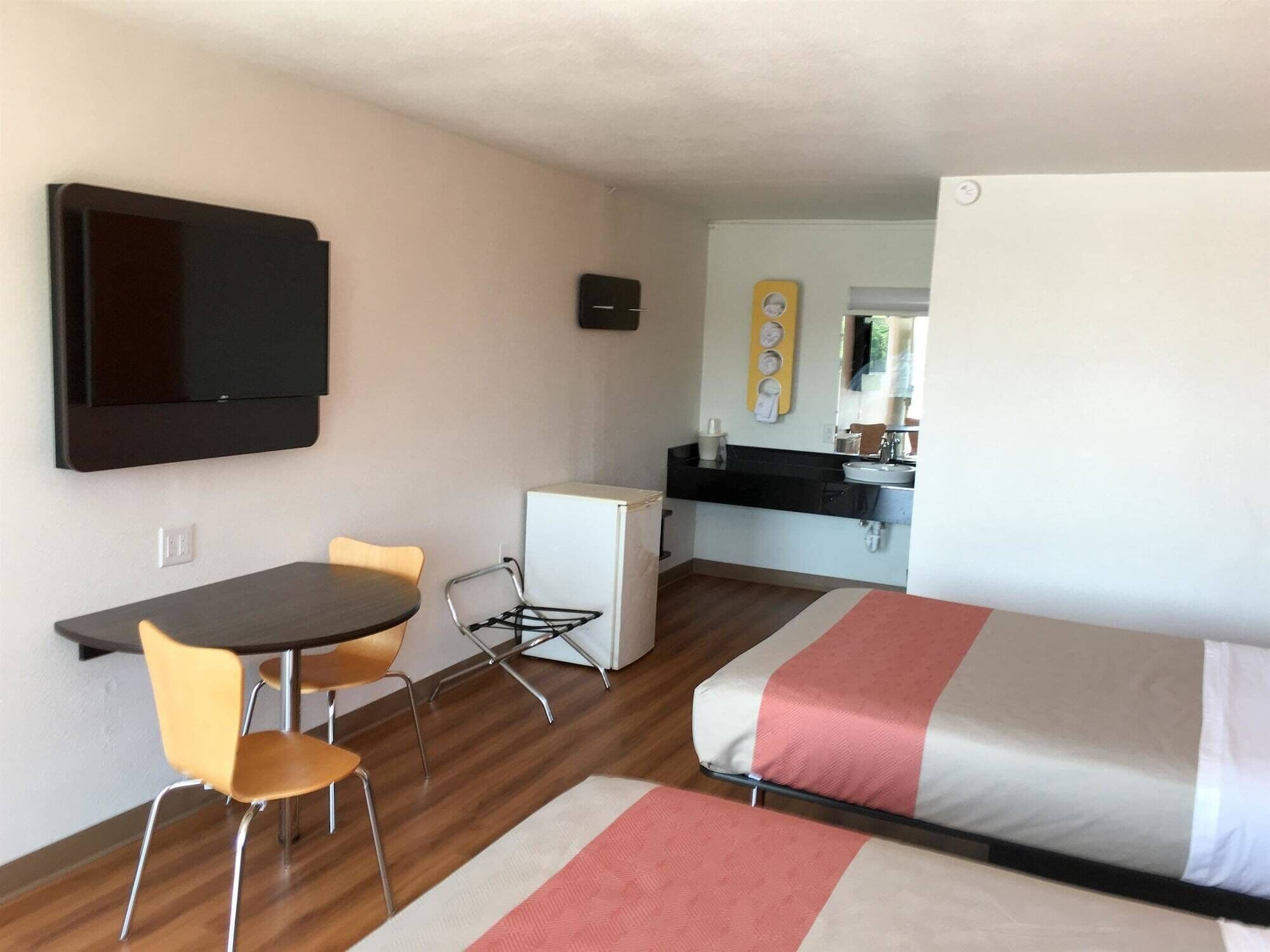 A compact room with a bed, a small kitchenette, a dining area with chairs, and a TV mounted on the wall.