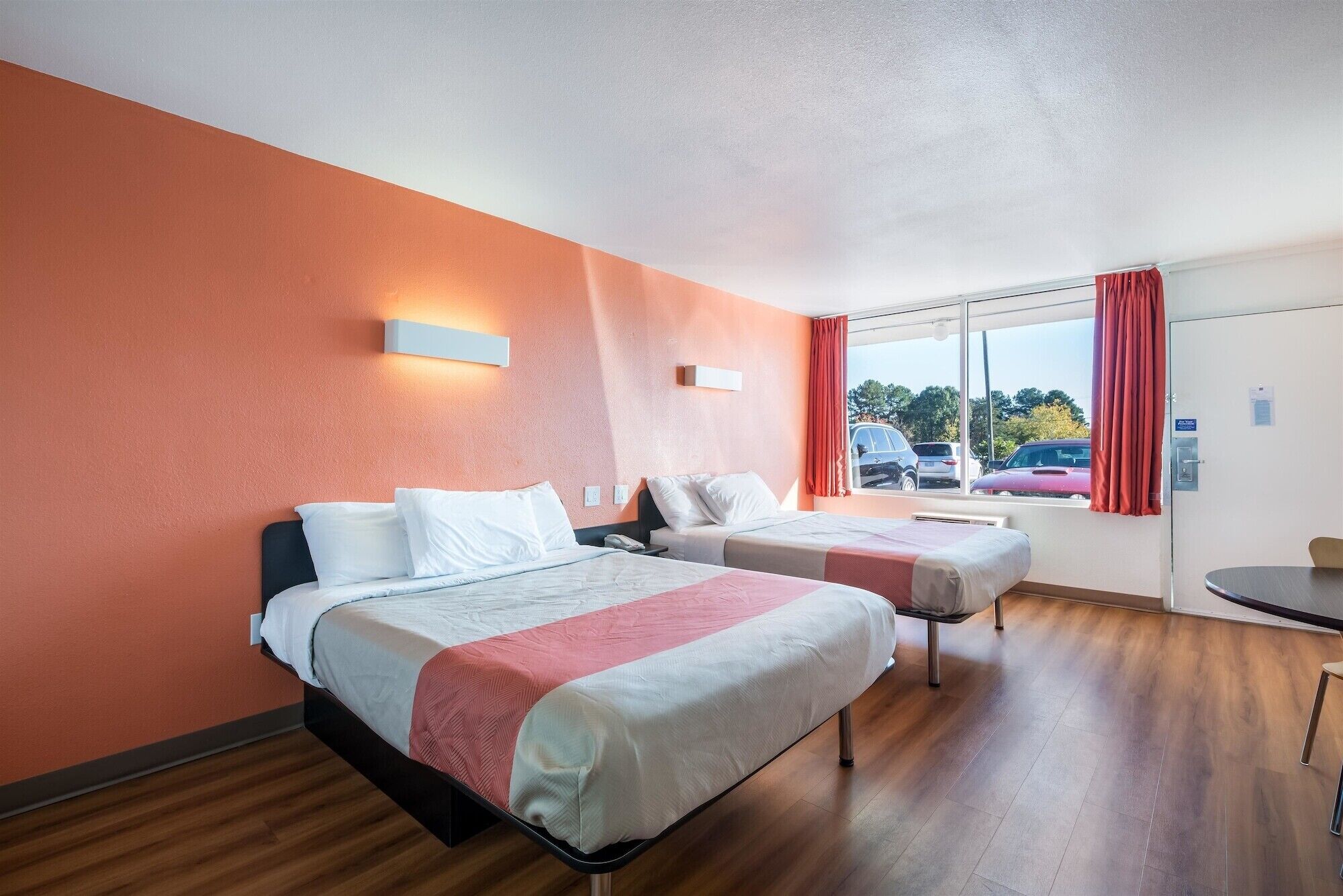 A hotel room with two beds, a window with a view of parked cars, and a small table.