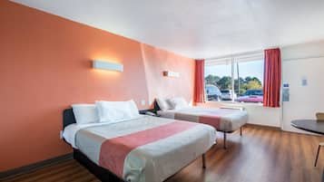 A hotel room with two beds, a window with a view of parked cars, and a small table.