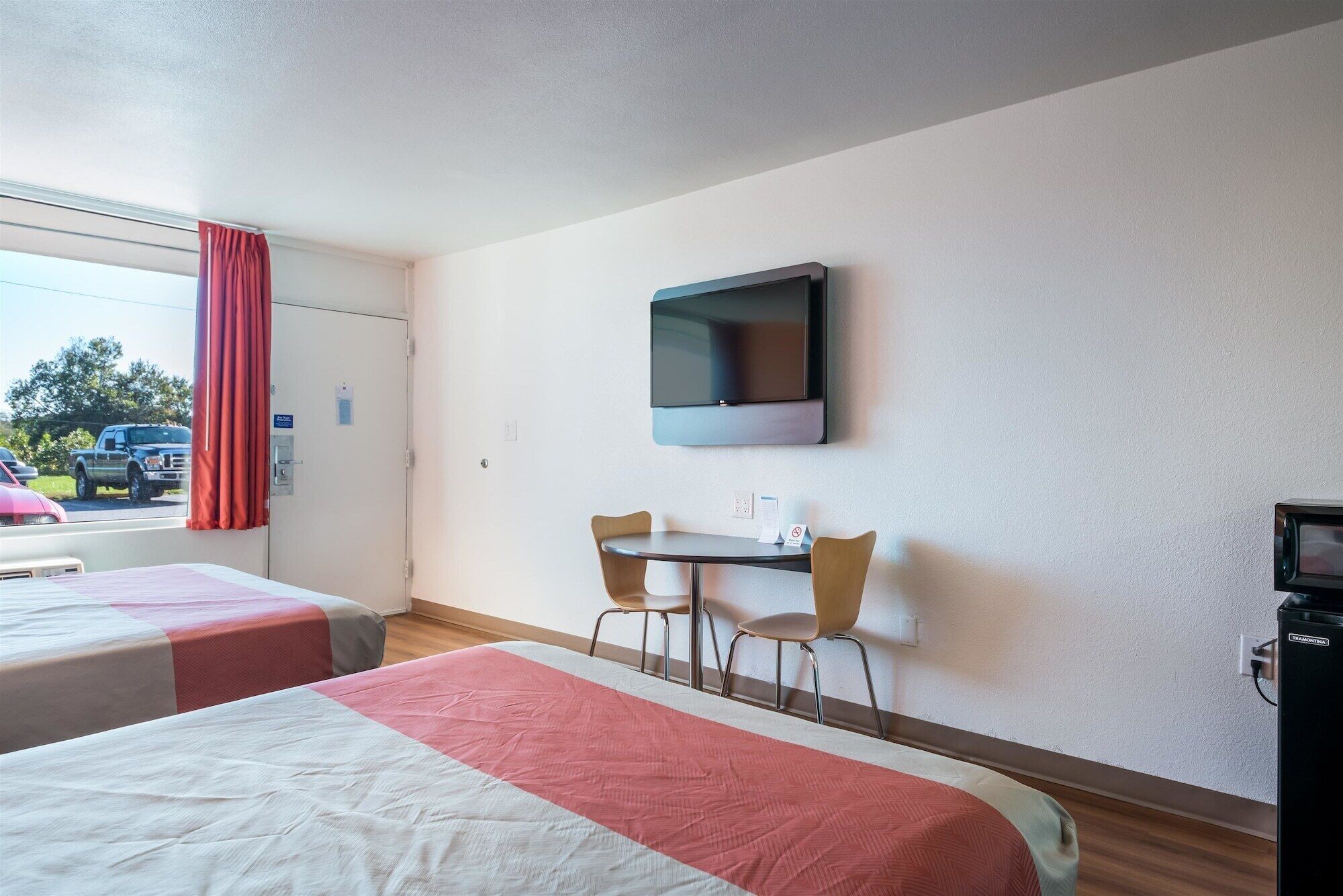 Habitación de hotel con dos camas, un televisor montado en la pared, una mesita con sillas y una ventana con cortina roja.