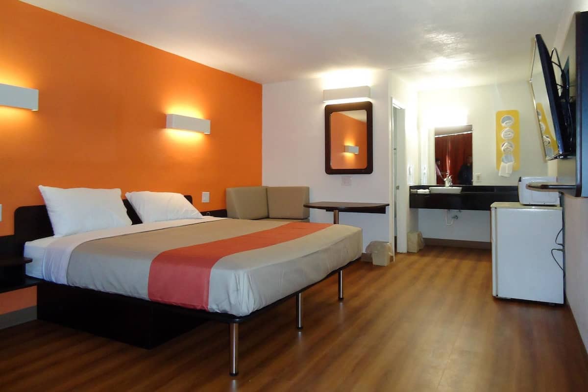 A hotel room with a bed, a small table, a chair, a desk, a mirror, and a refrigerator.