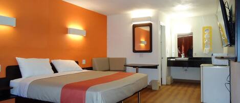 A hotel room with a bed, a small table, a chair, a desk, a mirror, and a refrigerator.