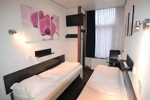 A hotel room with two single beds, a TV mounted on the wall, a window with curtains, and a small table with a chair. - Hotel Bienvenue (Rotterdam)