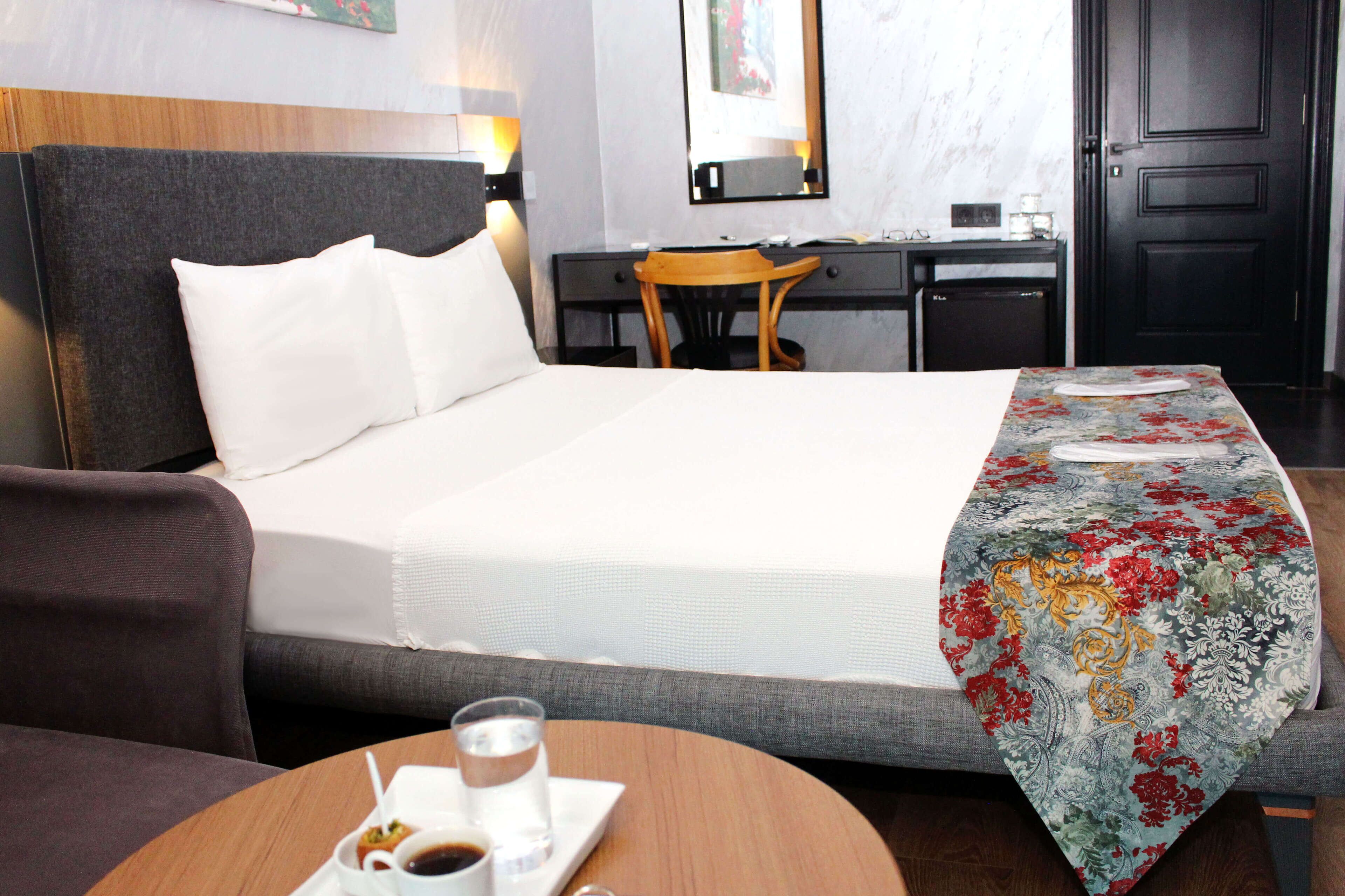 A hotel room with a bed, a desk, a chair, a table with a cup and a glass, and a mirror on the wall.