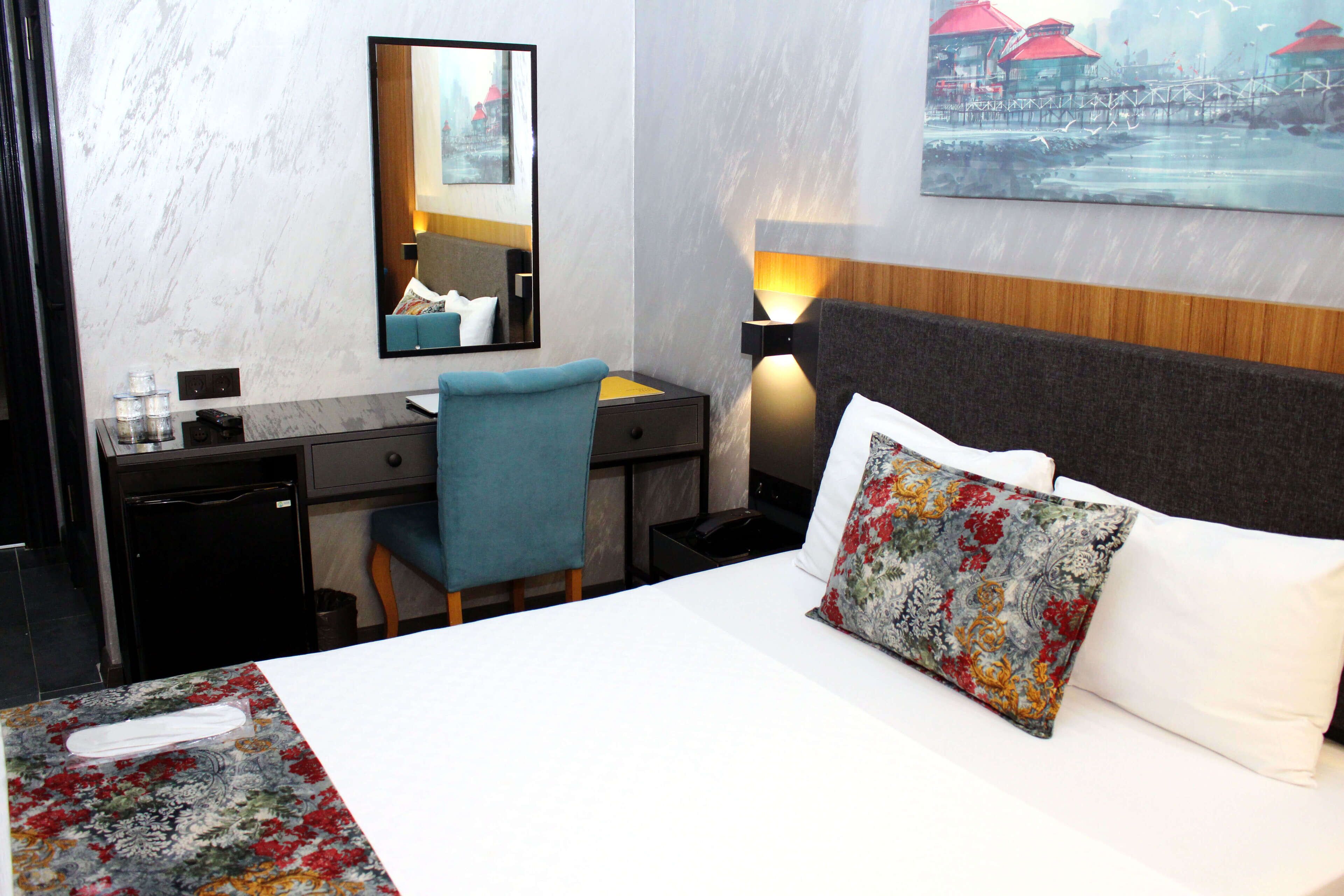 A hotel room with a bed, a desk, a chair, a mirror, and a painting on the wall.