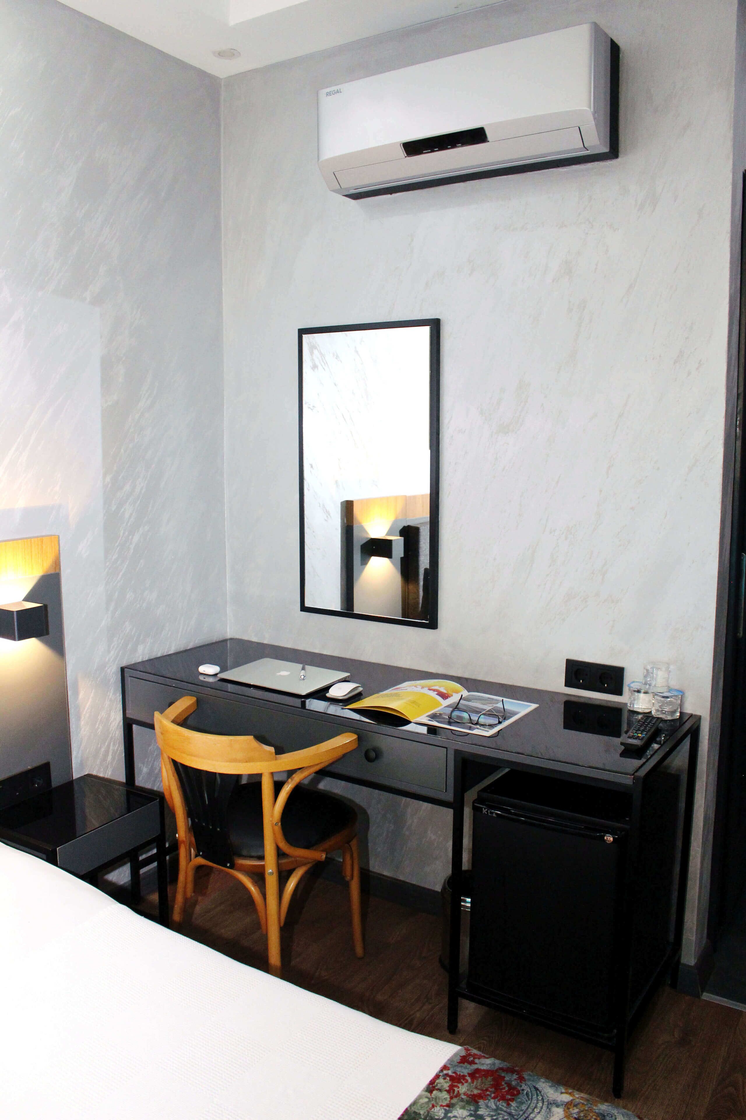 A hotel room with a desk, chair, and a small cabinet. There is a mirror on the wall, a lamp, and an air conditioning unit.