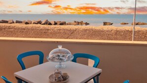 Una mesa con una cúpula de vidrio que contiene aperitivos, dos sillas azules y vista al mar con rocas al frente.
