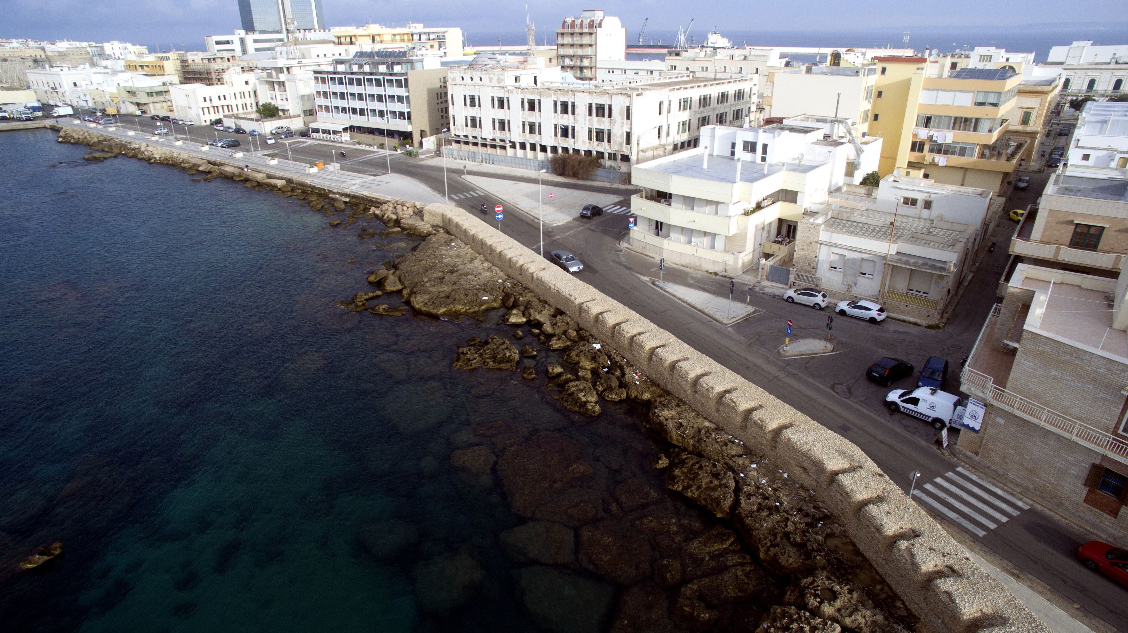 A coastal cityscape with a rocky shoreline, buildings, and a road with vehicles.