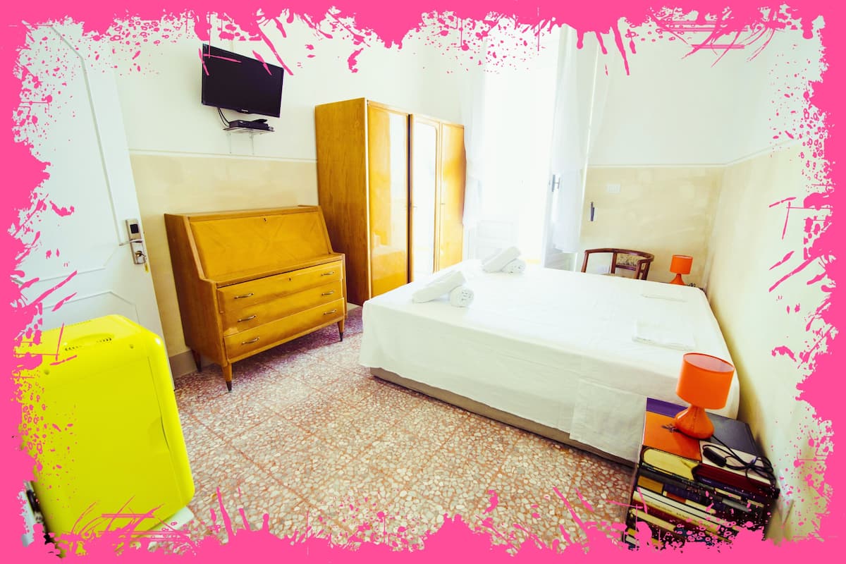 A bedroom with a bed, wooden wardrobe, a desk with a lamp, and a TV on the wall.