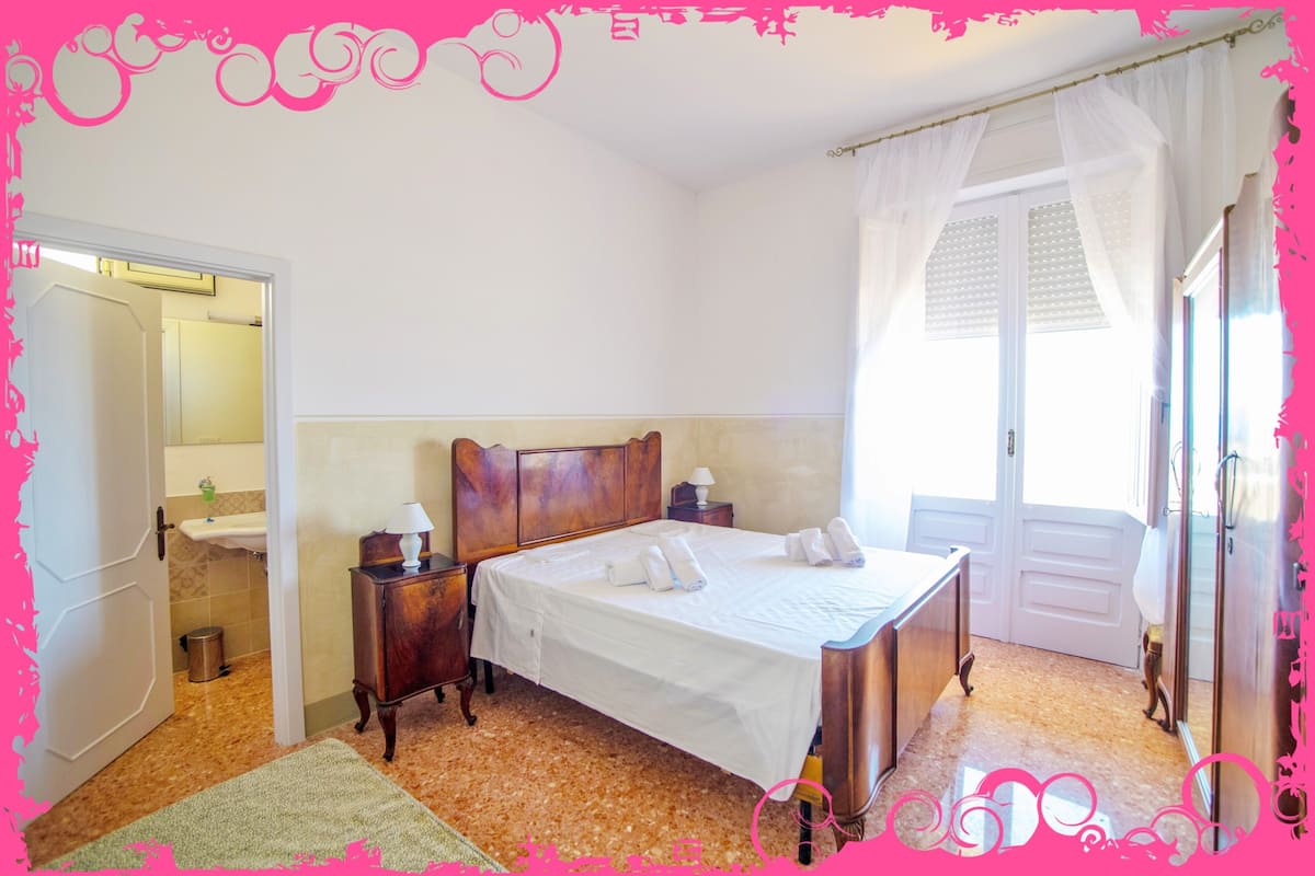 A bedroom with a bed, bedside tables, a nightstand, a window with curtains, and a door leading to a bathroom.