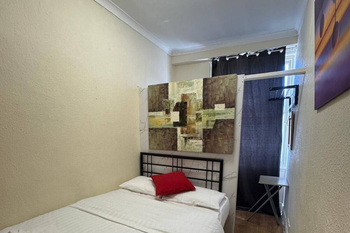 A small, clean room with a bed, a red pillow, and a painting on the wall.