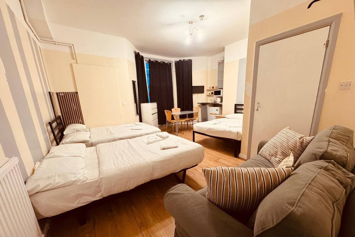 A compact hotel room with two single beds, a sofa, a small dining table, and a kitchenette.