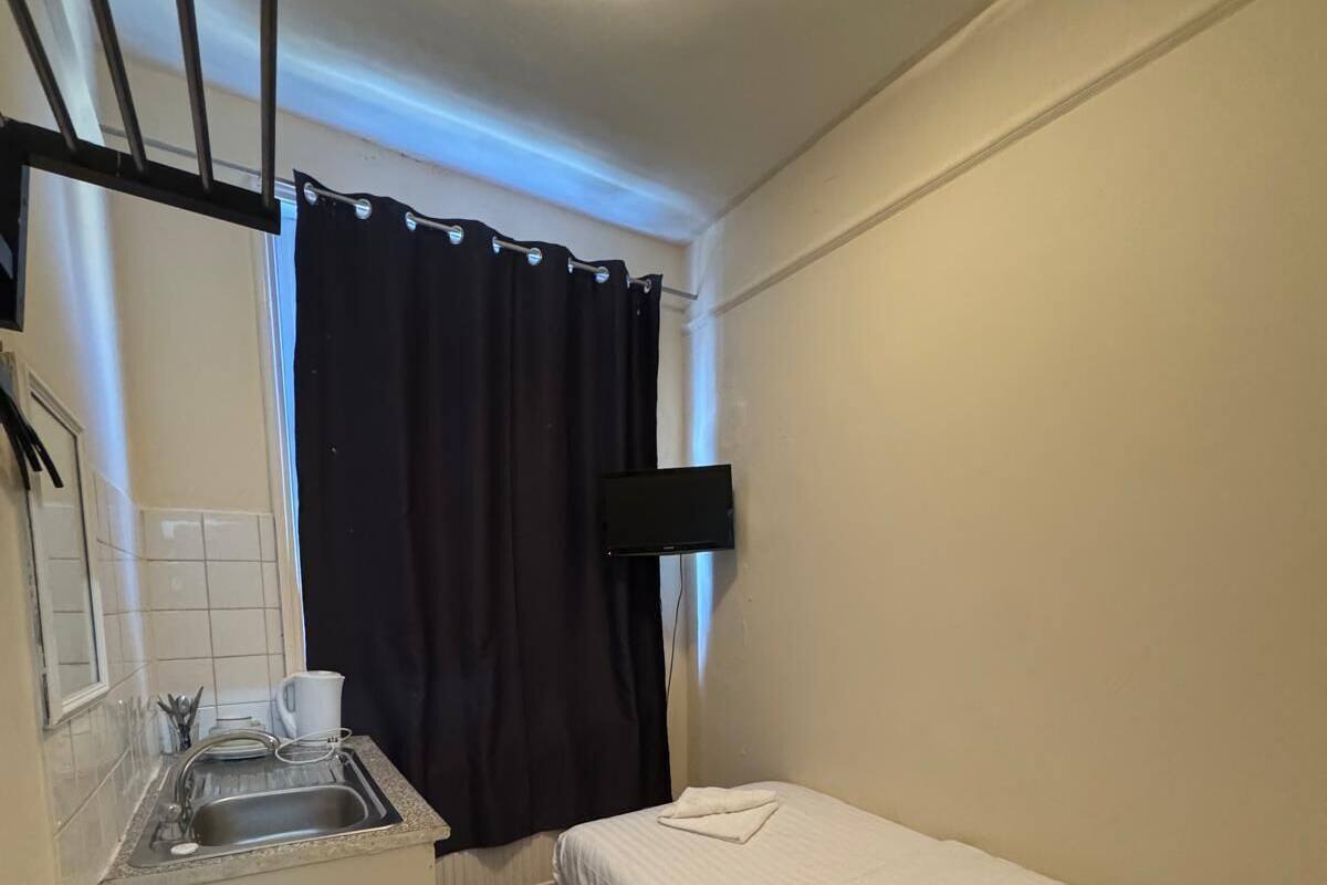 A compact room with a kitchenette, a single bed, a chair, and a black TV mounted on the wall.