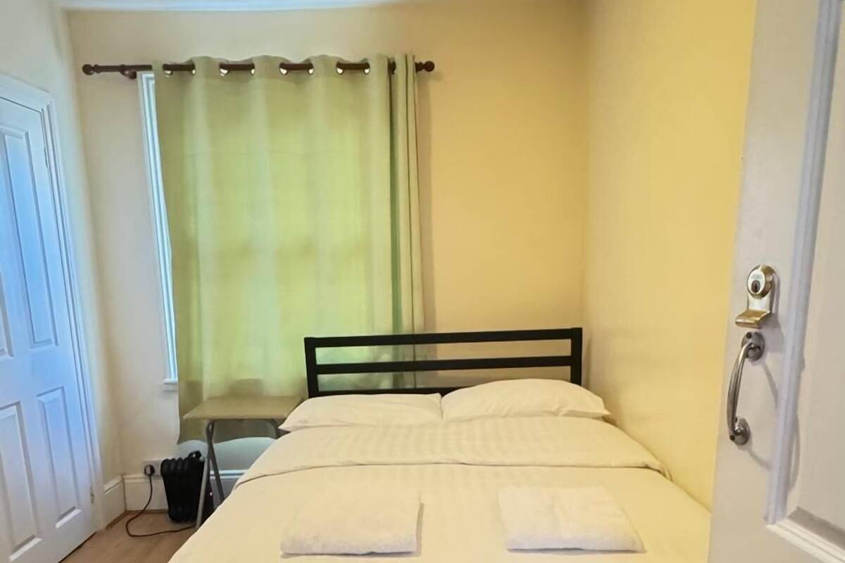 A small, neatly made bed in a room with wooden flooring, a window with curtains, and a door with a handle.