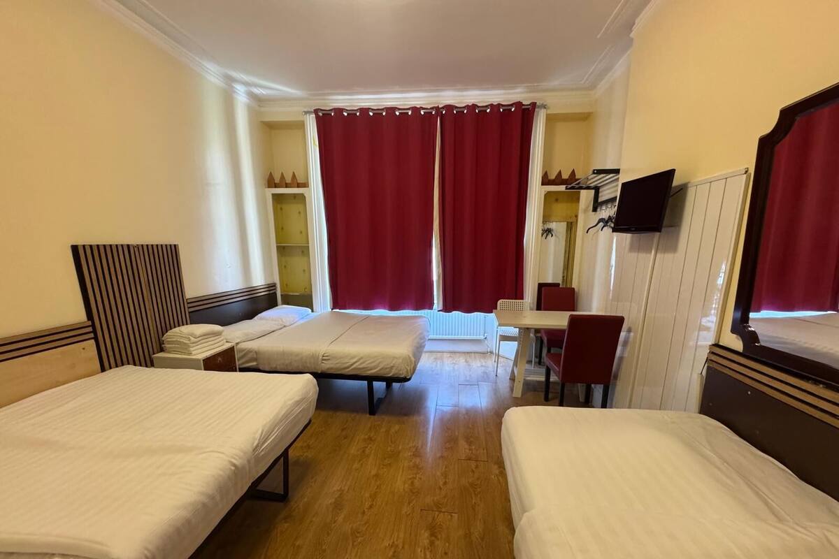 A hotel room with two single beds, a small table with chairs, a TV mounted on the wall, and a window with red curtains.