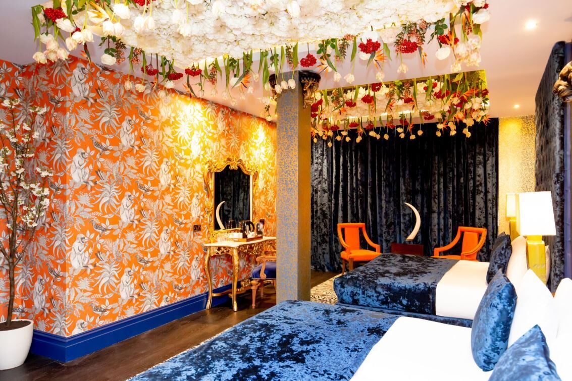 Una habitación con papel tapiz floral, un escritorio con sillas, una cama con ropa de cama azul y una araña con flores colgantes.