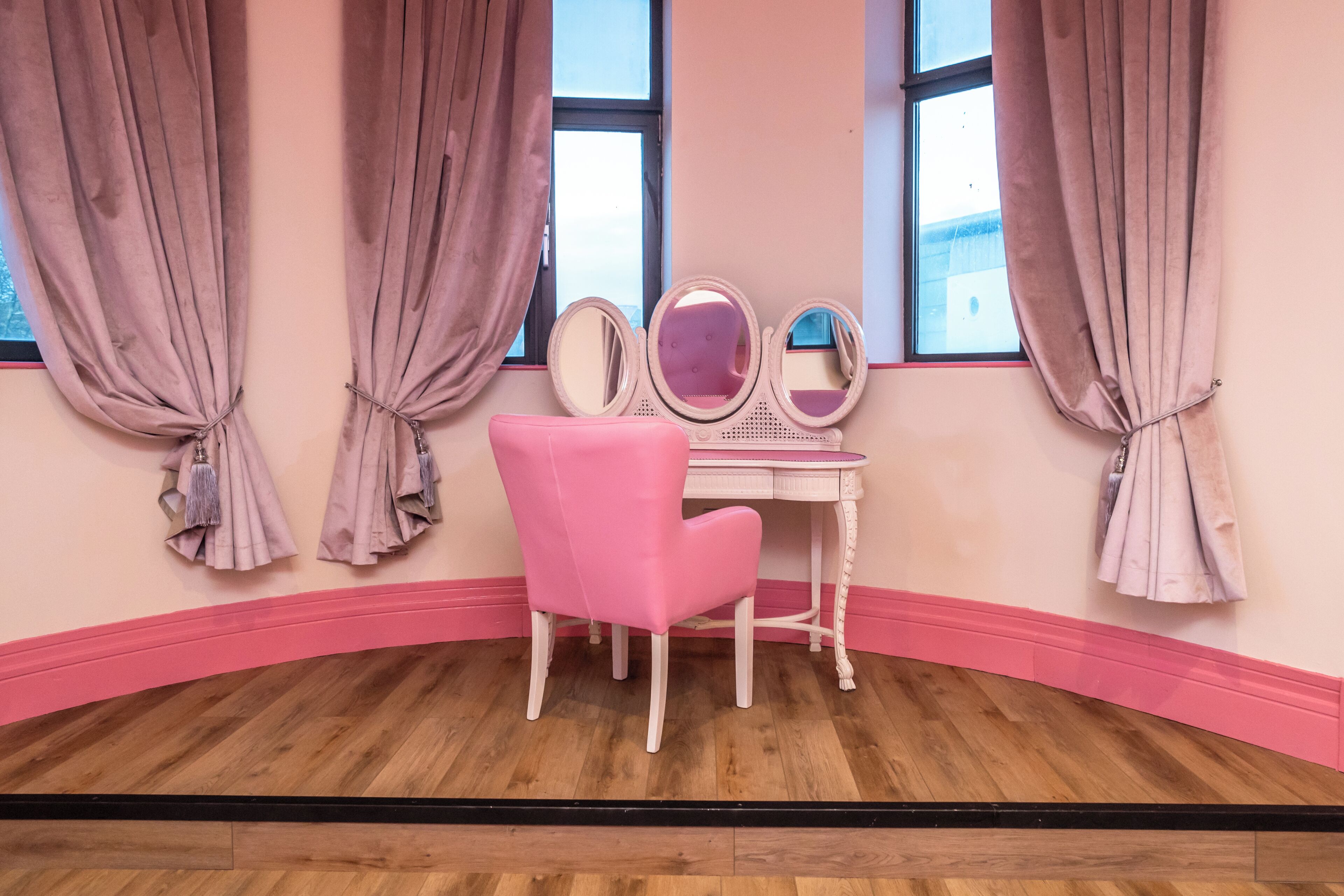 Une pièce avec une chaise rose, une coiffeuse avec des miroirs et des rideaux noués aux coins.