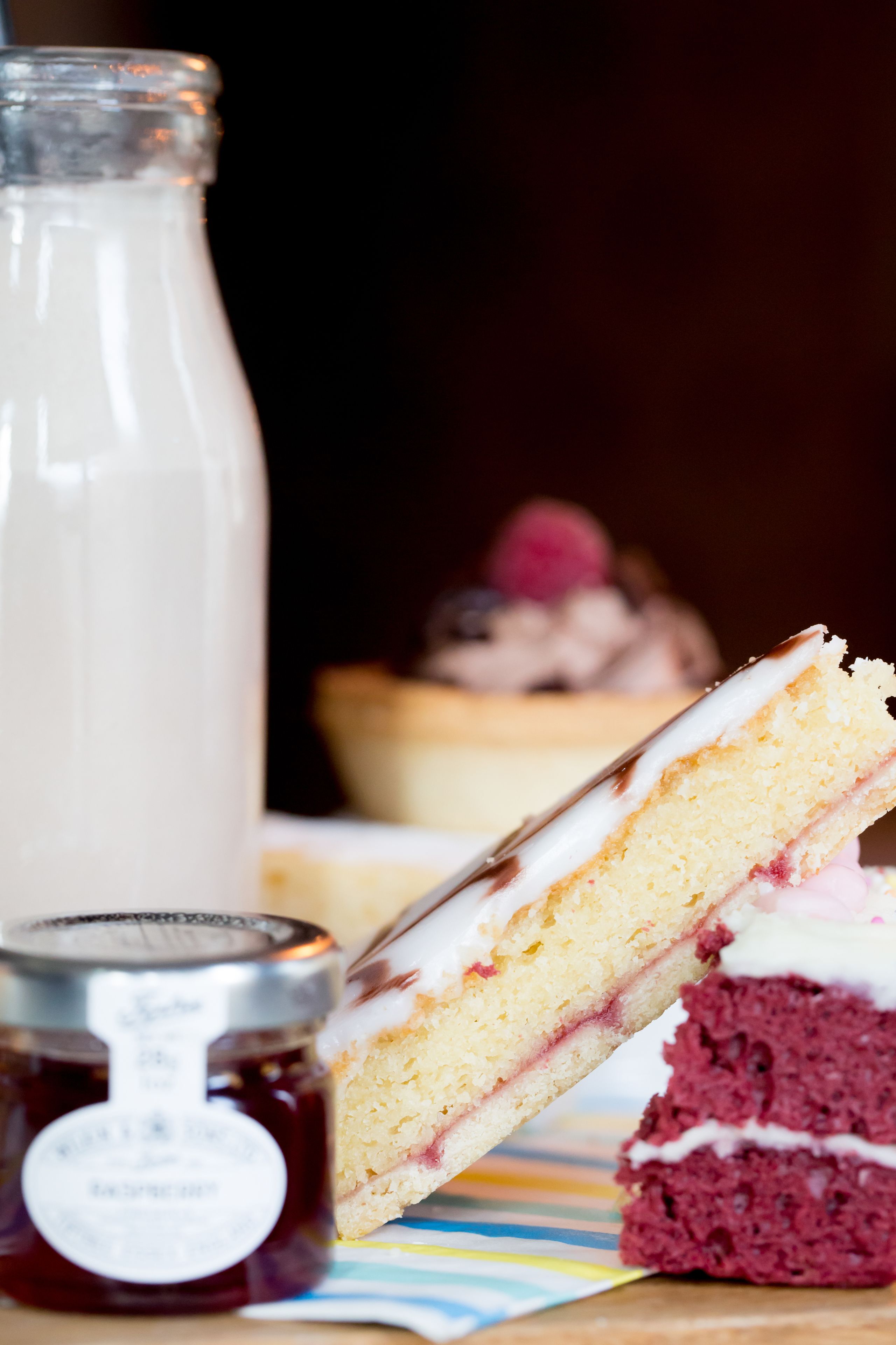 A slice of layered cake with white and red frosting, a jar of jam, and a glass bottle of milk on a striped tablecloth.