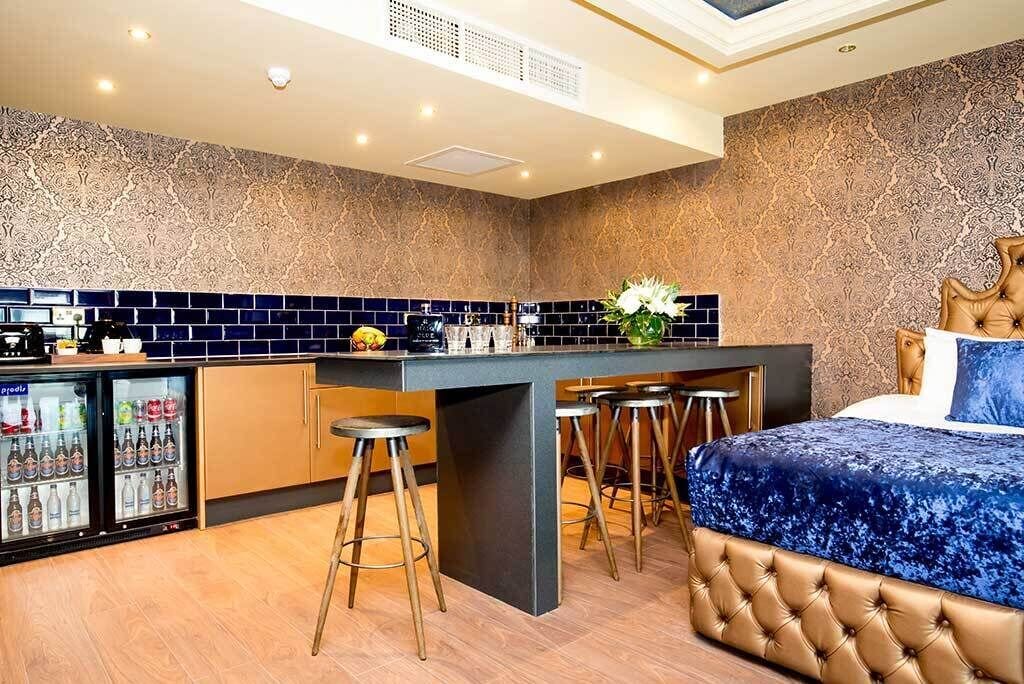A modern hotel room with a kitchenette, a bar area, and a large bed with a tufted headboard.