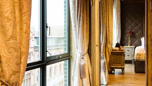 A room with a large ornate mirror, a wooden dresser, a bed, and a view of buildings through a large window.