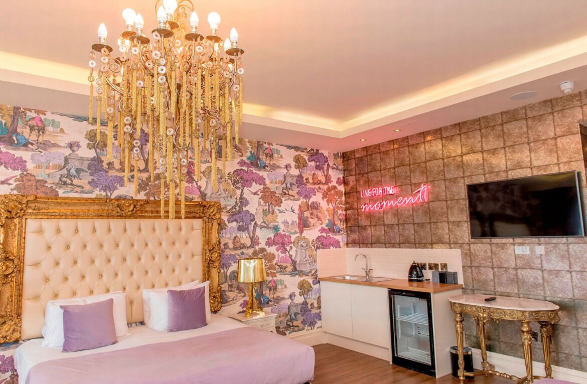 A bedroom with a large bed, a chandelier, a neon sign, a TV, a sink, and a small table.