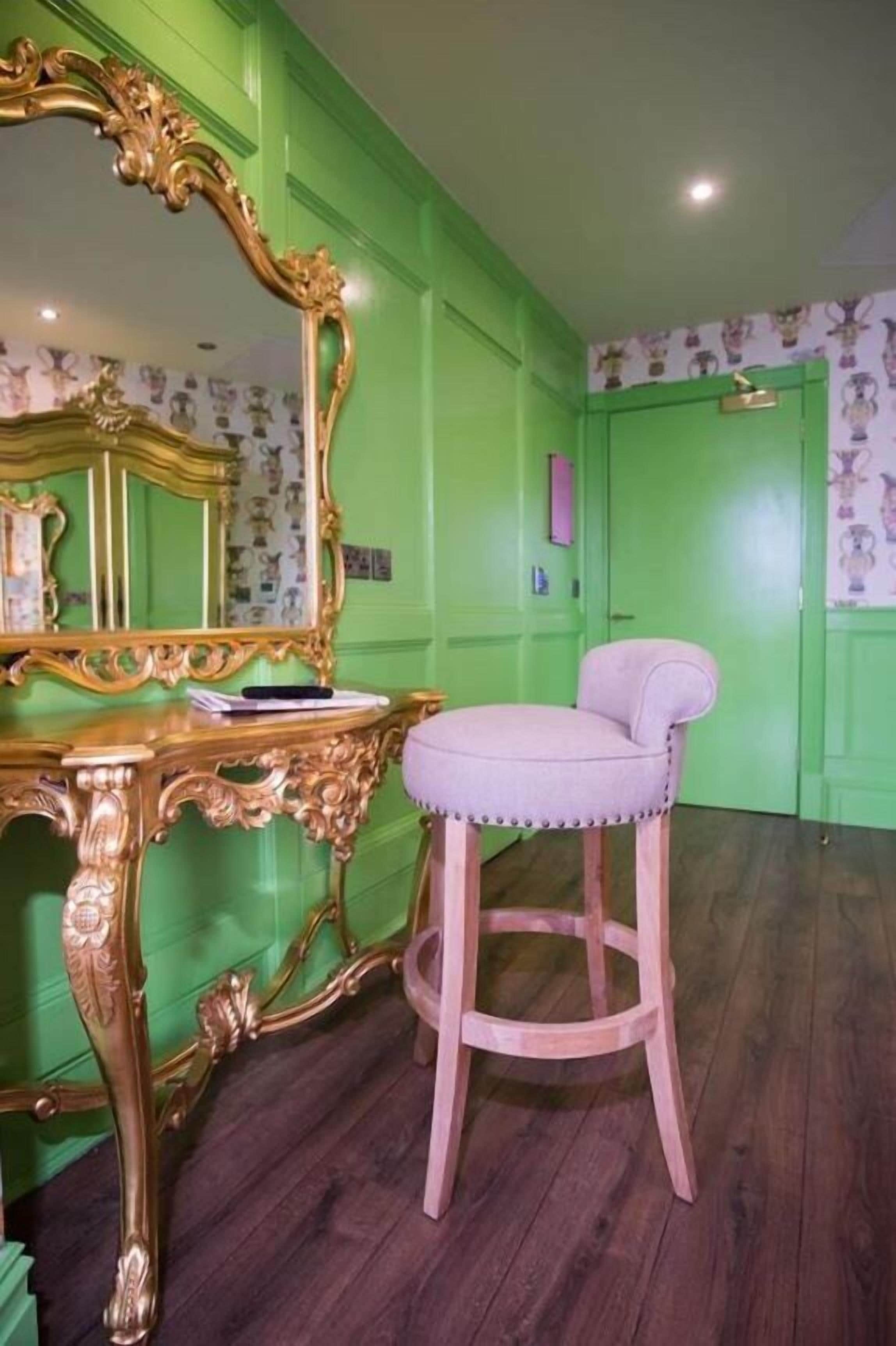 Une pièce avec un mur vert, un miroir encadré d’or, une coiffeuse en bois et une chaise rose.