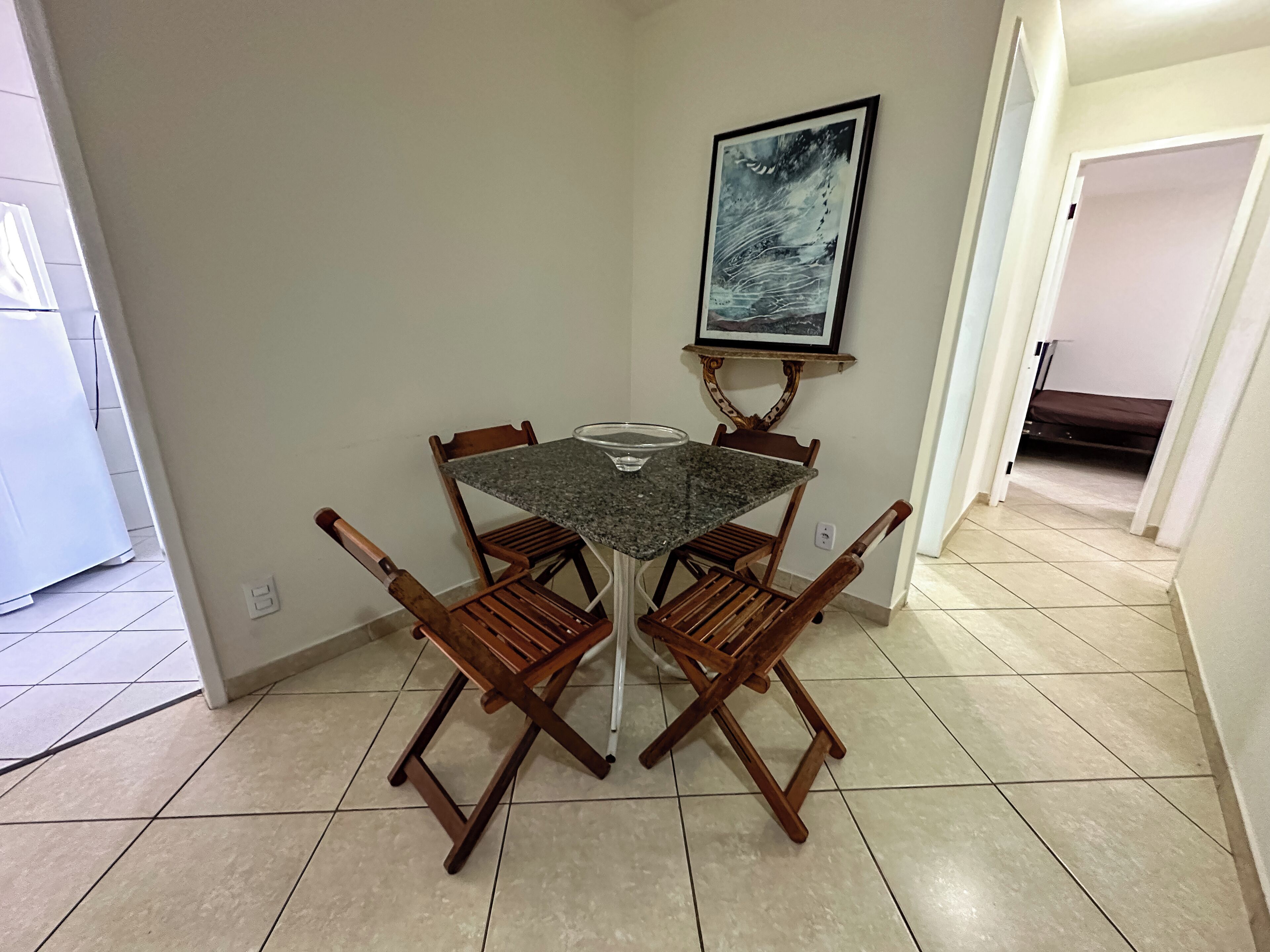 Un comedor con una mesa redonda, cuatro sillas de madera, un cuenco de vidrio y un cuadro en la pared.